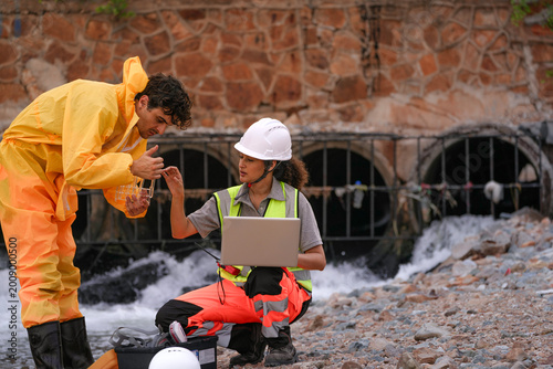 Obraz na plátně Two environmental field workers in protective gear conduct a water pollution inspection at a drainage outlet, collecting samples and analyzing data for contamination levels