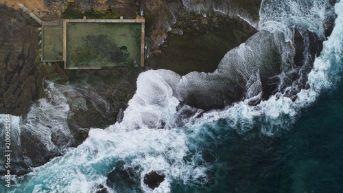 Aerial view of Newport Rock Pool with crashing white waves against dark coastal rocks and swimmers in the rectangular ocean pool in Sydney, New South Wales, Australia.