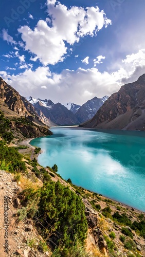 Stunning azure lake nestled between towering, rugged mountains under a partly cloudy sky. The landscape is vibrant and full of life
