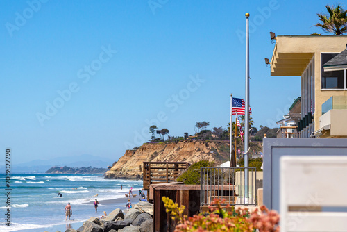 California beach with American flag and ocean waves. Scenic California beach coastline with people on the sand and rocky cliffs. Sunny day at California beach. Coastal landscape of California beach