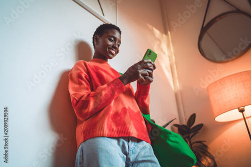 Foto Happy African American woman using mobile app, smartphone 5G connection, orange sweater, green shopper, digital nomad, authentic representation, inclusion, cozy indoor atmosphere, copy space