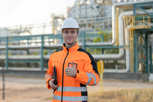 Photos A male engineer in an orange high-visibility jacket and white helmet gives a thumbs-up at an industrial plant with pipelines in the background