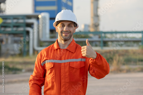 Canvas Print A construction worker in an orange safety uniform and white helmet gives a thumbs-up with an industrial site in the background