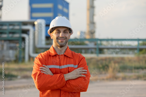 Fotografi A male worker in an orange safety uniform and white helmet stands confidently at an industrial site with pipelines and structures in the background