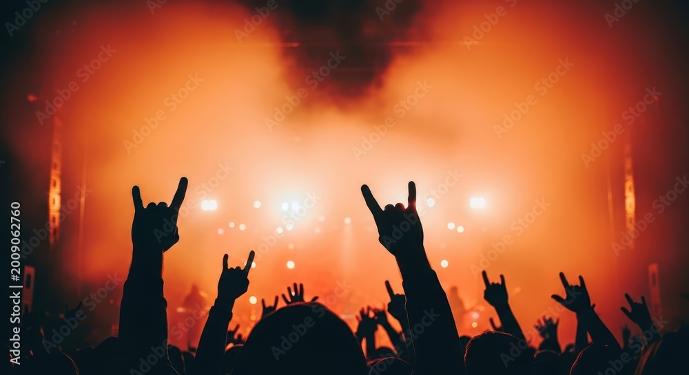 Fototapeta premium Silhouetted hands make rock gesture signs in front of a brightly lit stage with hazy atmosphere.
