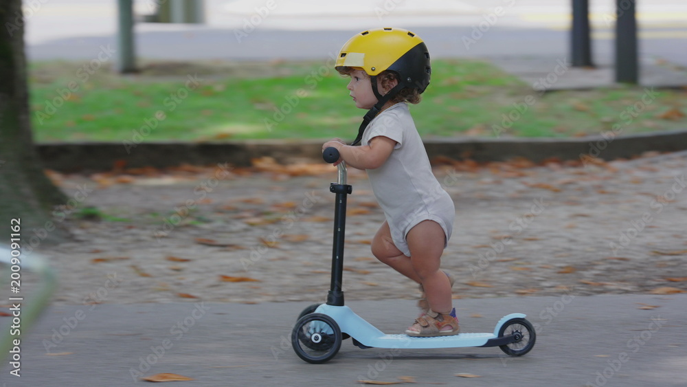Fototapeta premium Toddler wearing yellow helmet riding blue scooter in park pathway, captured in side profile, focused and balanced while gliding forward during playful outdoor moment