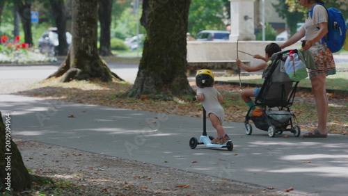 Toddler riding scooter on sunny park pathway wearing yellow helmet, distant view with parent and stroller ahead, playful and curious outdoor childhood activity