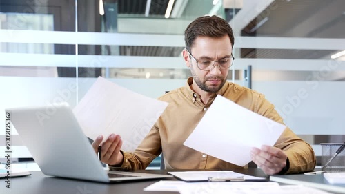 Confused businessman reviewing financial documents at desk in modern office, struggling to understand poor results. Frustration, analysis difficulty and concern during stressful business workflow.
