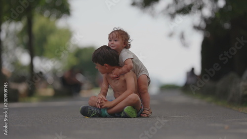 Fotografía Toddler hugging older brother from behind while sitting together on park pathway