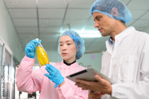 Food quality inspectors checking bottled fruit juice on production line in beverage factory, using tablet for monitoring and quality assurance.