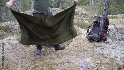 A male hiker packing sleeping mat on rocky mossy ground in a northern forest, with a backpack nearby during an outdoor camping trip.