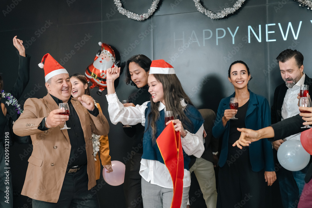 Fototapeta premium New year party celebration with smiling adult people, festive hat, wine glass, balloon, and holiday decoration in indoor office event