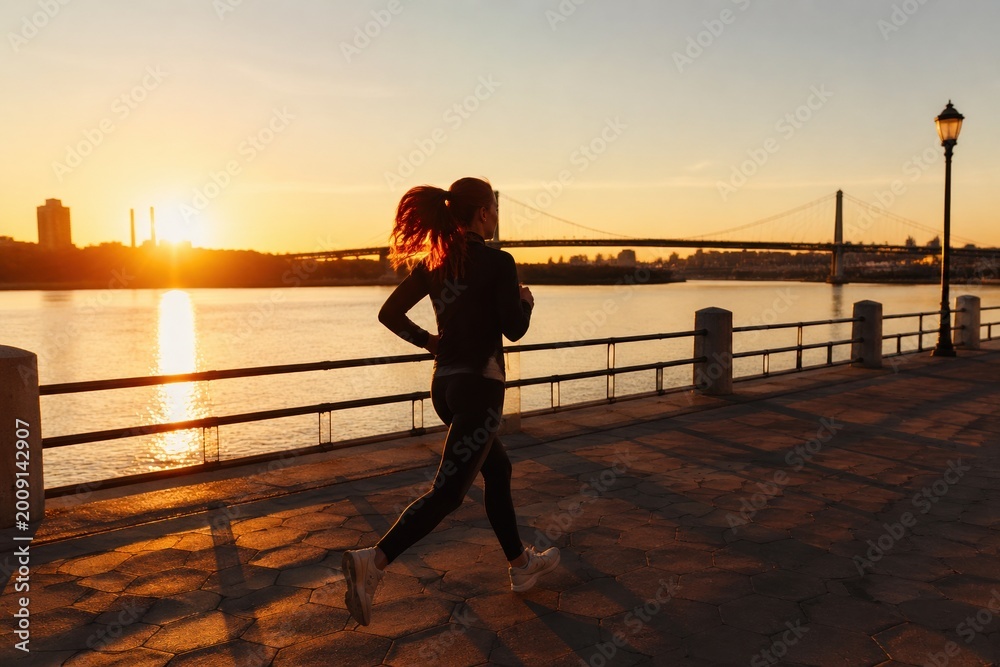 Fototapeta premium Red haired woman running on city embankment at sunset. The scene captures an energetic yet peaceful mood with golden sunlight. Ideal for fitness, health, and lifestyle marketing materials.