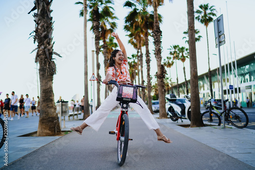 Laughing young Black woman cycling with arm raised in joy, embracing digital age mobility, feminine empowerment and vibrant lifestyle through modern shared transport.