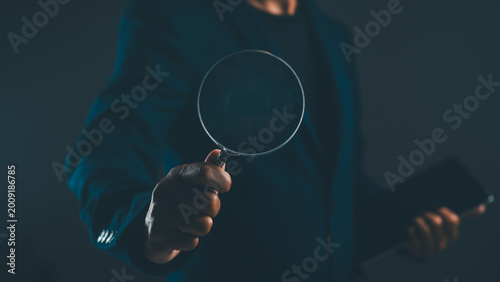 Fotografie A person in dark clothing holding a magnifying glass, focusing on an object in a dimly lit environment
