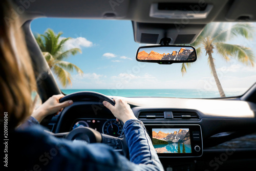 View from the back seat inside car, showing woman’s hands on the steering wheel. Through the front windshield, open road leads toward scenic landscape of beach and mountains. Travel concept.