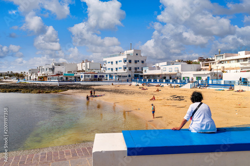 Urban beach of Corralejo with people, calm water and white houses along promenade under blue sky with clouds.