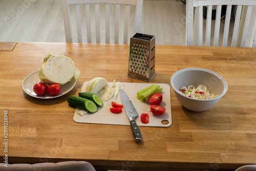 Simple home cooking. Wooden table with fresh vegetables, sliced cabbage, halved tomatoes, cucumber, cutting board knife. Preparing nutritious midday meals. Creating nourishing. Assembling hearty lunch