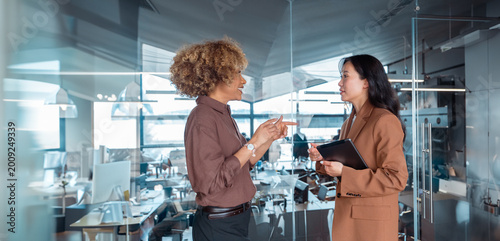 Two diverse professional women having business discussion standing in modern office, holding tablet, teamwork and business communication. Business people communicating at work. Web banner, copy space