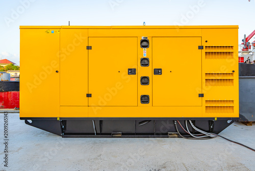 Large industrial diesel generator in yellow soundproof canopy outdoors on a concrete platform under a clear sky