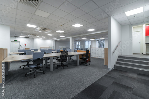 The scene shows an empty office with desks arranged in an open layout. Natural light comes through large windows. There are chairs at each desk, and some steps lead to another area