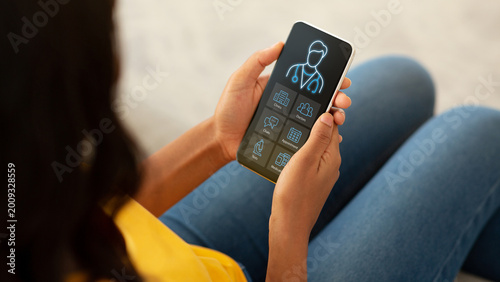 Telemedicine Concept. Closeup of black woman patient holding mobile phone with online medical services on screen, register online appointment to physician, home interior, cropped