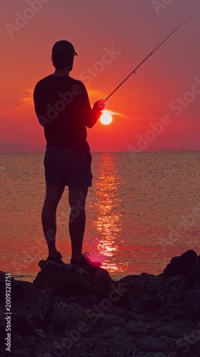 Vertical video. Man Fishing from Rocks Facing Sunset Over Sea Wide Angle Silhouette Outdoor Adventure Coastal Angling Scene