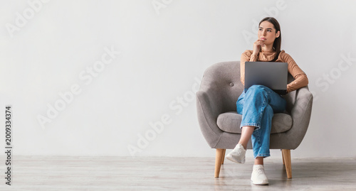 Foto Thoughtful millennial lady with laptop pc sitting in armchair, looking at empty space against white studio wall, banner design