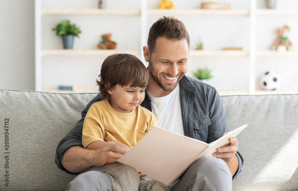 Naklejka premium Weekend activities with toddlers. Cheerful carying daddy reading book to little son, baby boy enjoying fairy tale on dad's knees, sitting on sofa at home