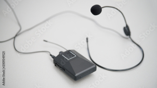 Black radio microphone system with a portable transmitter unit resting on a bright desk. Perfect template for public speaking, fitness instruction, and media event designs. Photo