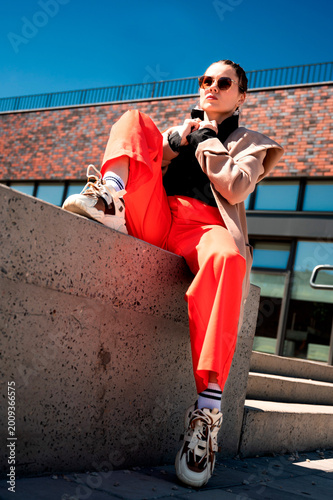 Fotografija A photograph of a fashionable young woman in a beige jacket, black shirt, and red pants, posing confidently on a sunlit city street