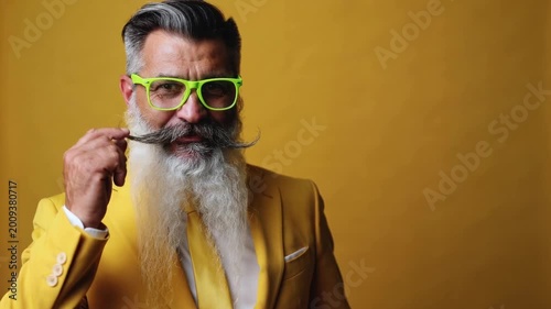 Mature man in yellow suit and neon green glasses touching mustache against yellow background medium shot