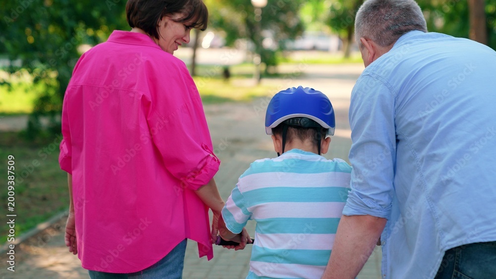 Naklejka premium Child son cyclist with parents in city park. Happy family, child in helmet, mother father playing with bicycle outdoors in summer. Family day out. Dad mom teach son to ride bike in park. People park