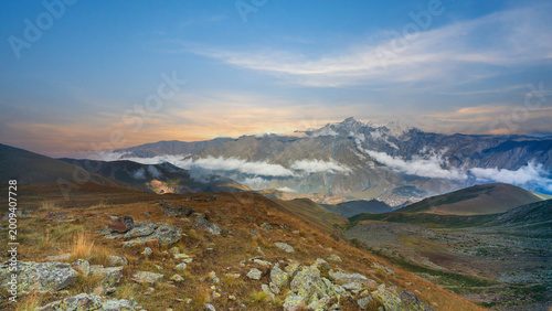 Mountain landscape above Kazbegi in Georgia with rocky foreground, low clouds, and distant snowy peaks at sunset.
