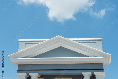 Tablou pe pânză A peaked roof, a portico, at the entryway of a vintage pale blue building with wide decorative triangular moulding over the doors