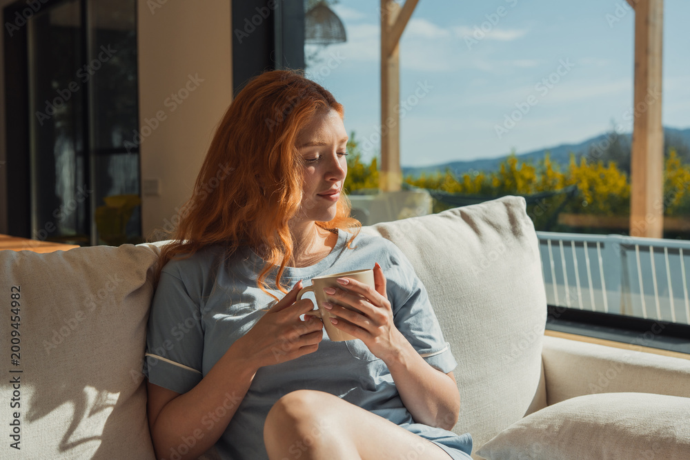 Naklejka premium A red-haired woman sits on a light sofa holding a mug, enjoying warm morning sunlight through large windows onto a terrace and distant hills, conveying a calm, cozy and reflective at-home moment.