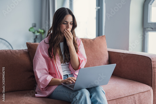 Cuadro en lienzo Young woman sits on a couch with a laptop, looking shocked and worried while reading something on screen; conveys stress, anxiety, remote work and unexpected online news at home
