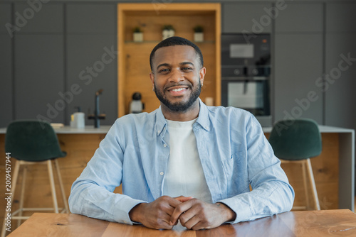 Photography A relaxed young man smiles at the camera while seated at a wooden kitchen table in a modern home