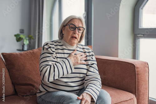 Konstfotografi An older woman sitting on a couch holds her chest and looks distressed, suggesting chest pain, shortness of breath or medical emergency at home