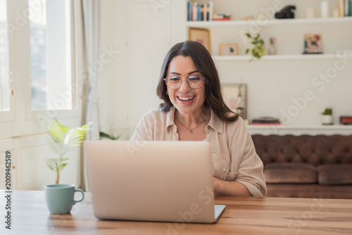Smiling woman using a laptop for a video call in a bright, cozy home office, engaged in remote work or an online meeting, conveying warmth, focus and connection.