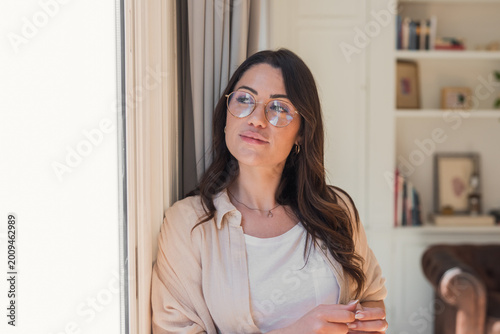 Obraz na płótnie A thoughtful young woman in glasses gazes out a window in a bright, cozy living room, wearing casual clothing and a soft smile that conveys calm, reflection and quiet confidence