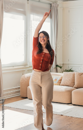 A smiling young woman dances joyfully in a bright, airy living room, wearing casual clothes and glasses while enjoying a relaxed, confident moment at home surrounded by cozy decor.