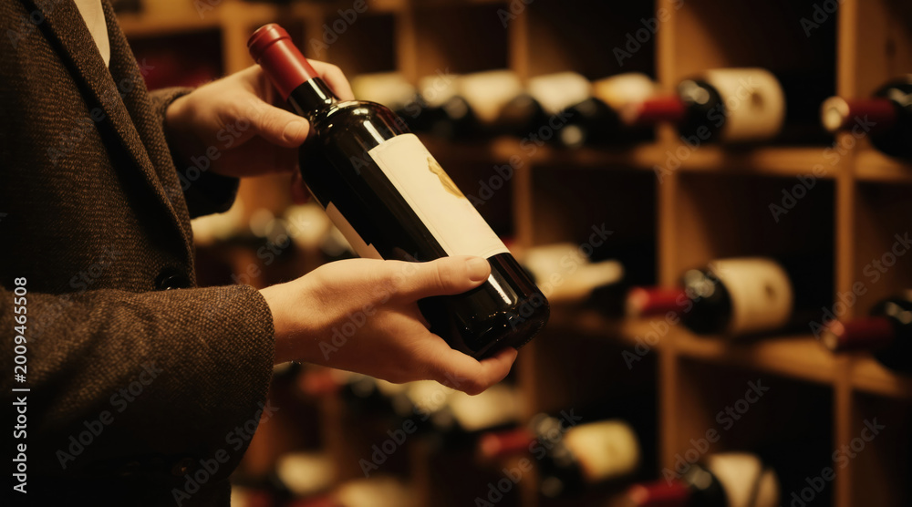 Naklejka premium Man picks wine bottle from wooden shelf in dark cellar with rows of alcohol drinks. Person examines beverage for tasting at winery or bar. Expertise for connoisseur.