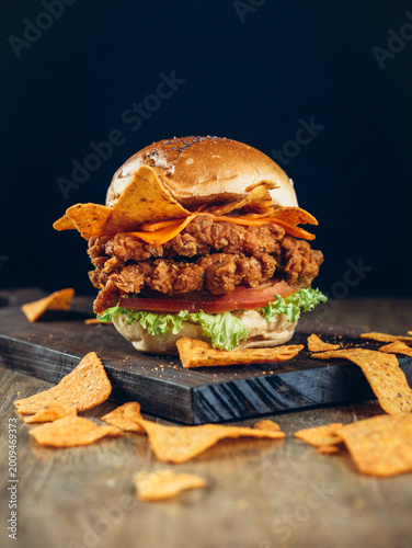 Crispy fried chicken burger with cheese, tortilla chips, lettuce, and tomato on a toasted bun, shot on a dark background.
