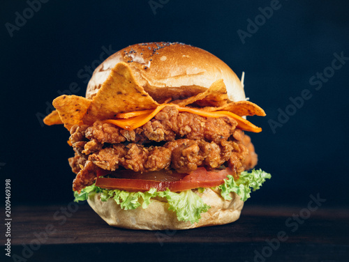 Crispy fried chicken burger with cheese, tortilla chips, lettuce, and tomato on a toasted bun, shot on a dark background.
