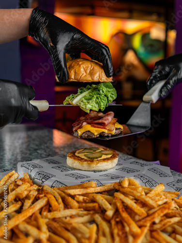 Gourmet cheeseburger being assembled with bacon, lettuce, and pickles, served with seasoned fries in a vibrant kitchen setting.