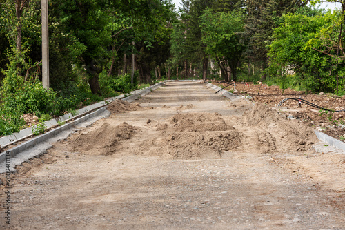 Road construction and landscaping works in a city park. The image shows newly installed concrete curbs, piles of sand, and a leveled dirt surface prepared for paving or asphalt.