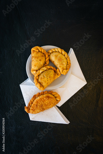 Vista cenital de empanadas fritas dispuestas sobre un plato blanco y servilletas, con un fuerte contraste sobre fondo de mesa negro rústico.