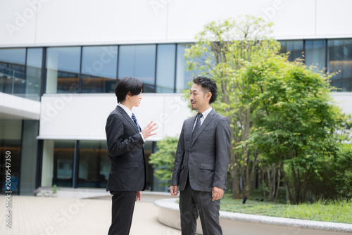 屋外のビルの前で信頼感あるスーツの営業チームのコミュニケーション　部下と上司が会話する話すシーン全身