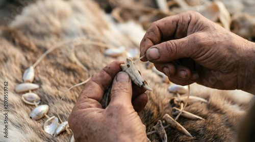 Close-up of weathered hands crafting a primitive tribal necklace with animal teeth and seashells over a textured animal fur background in a workshop.